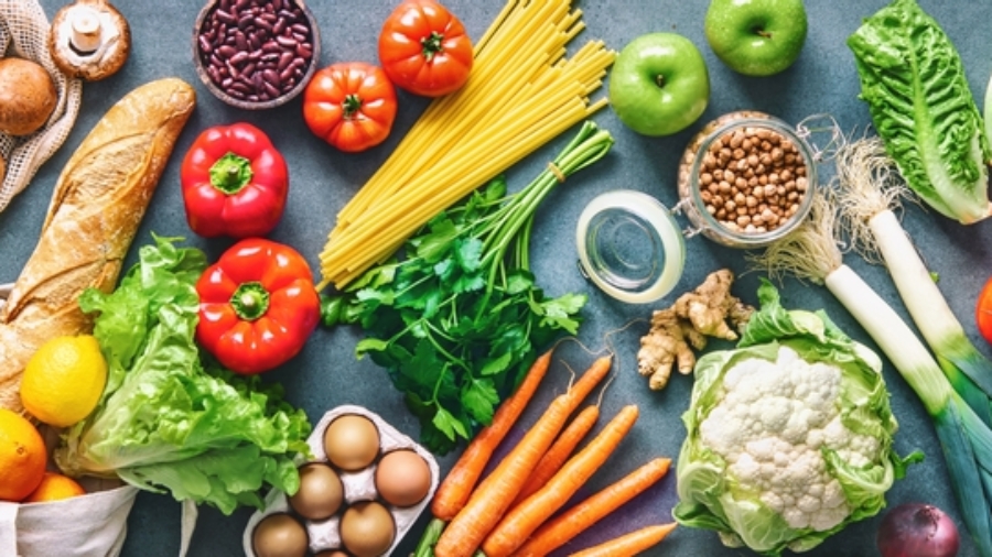 A variety of fresh foods including vegetables, fruits, bread, eggs, pasta, beans, olive oil, and milk are arranged on a table, showcasing healthy ingredients for balanced meals.