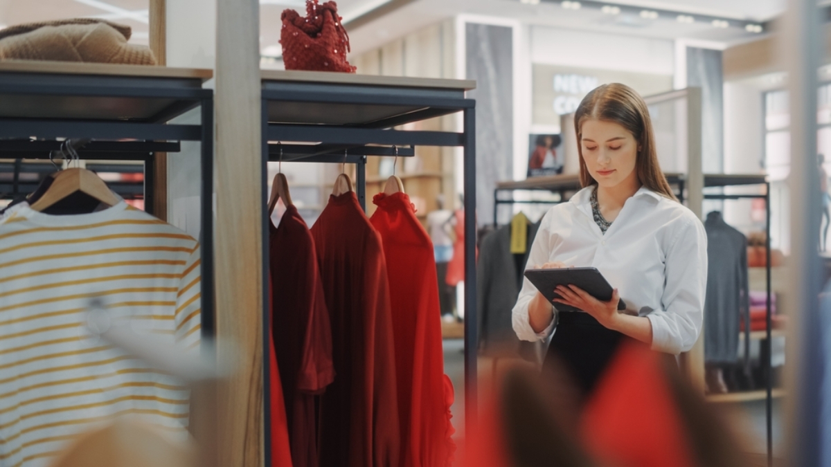 A woman in a white blouse stands in a modern, well-lit clothing store, holding a tablet with an OMS interface. Red dresses and a striped shirt hang on racks around her as she manages the store’s inventory seamlessly.