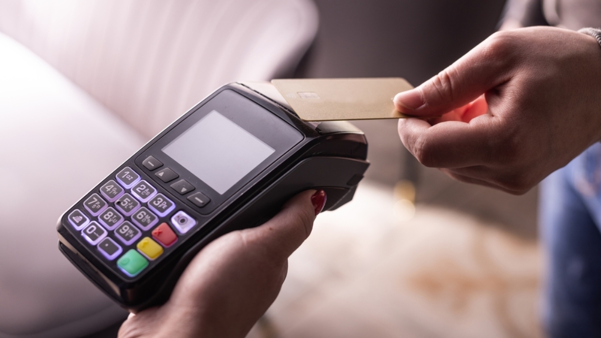 A person holds a payment terminal while another taps a gold credit card, showcasing how contactless payments can help automate everyday transactions.