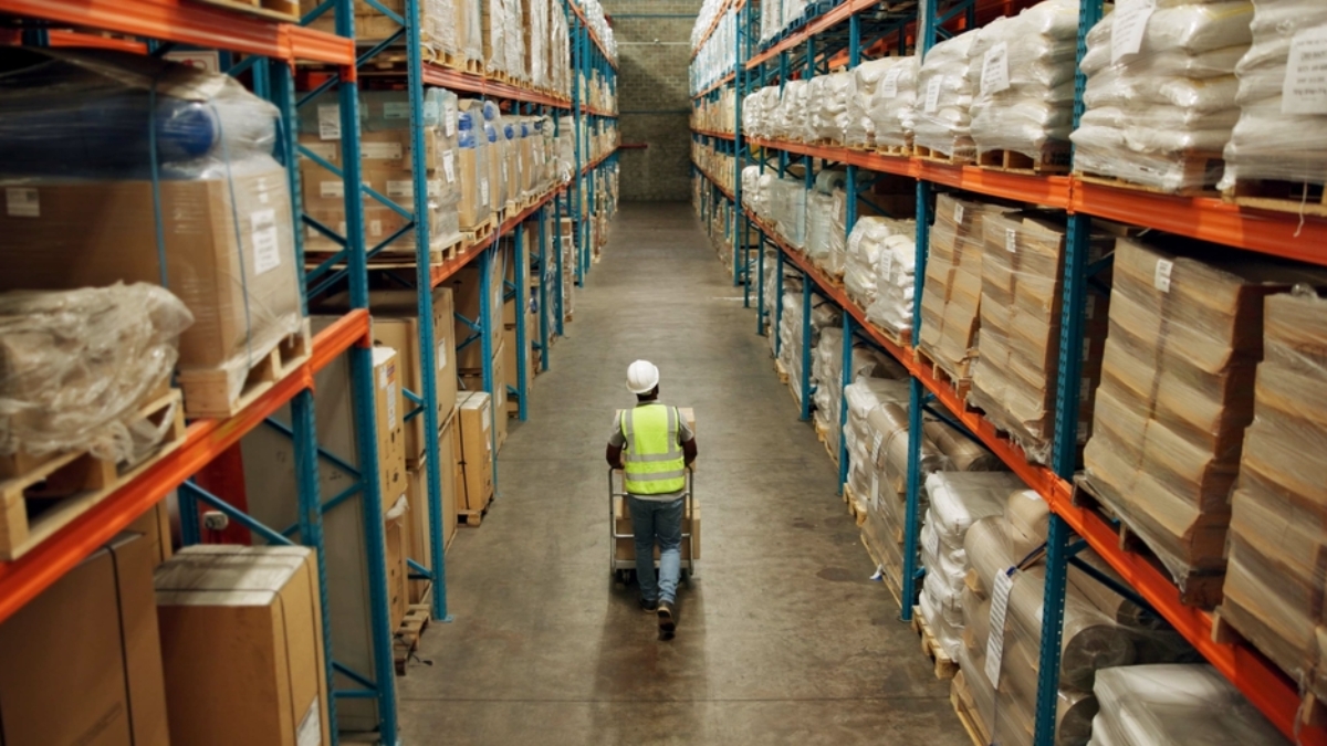 A warehouse worker in a reflective vest and hard hat pushes a cart down a wide aisle between tall shelves filled with stacked boxes and wrapped pallets.