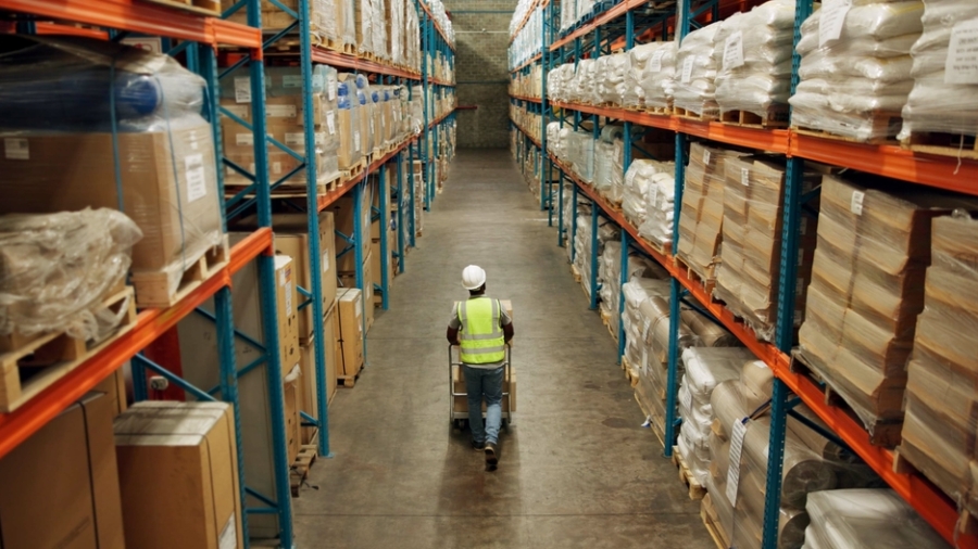 A warehouse worker in a reflective vest and hard hat pushes a cart down a wide aisle between tall shelves filled with stacked boxes and wrapped pallets.