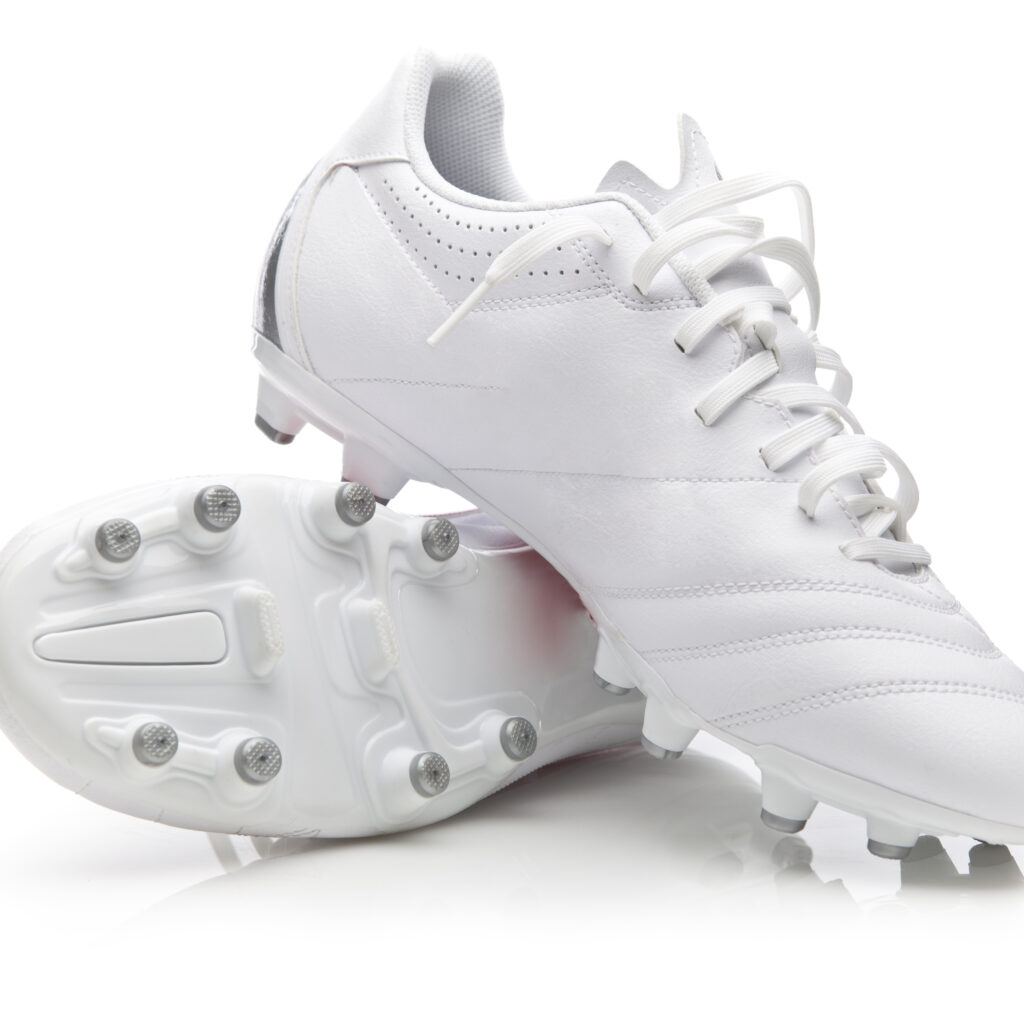 A pair of clean, white soccer cleats with laces, one shoe laying on its side showing the sole and studs, and the other upright, both on a white background.