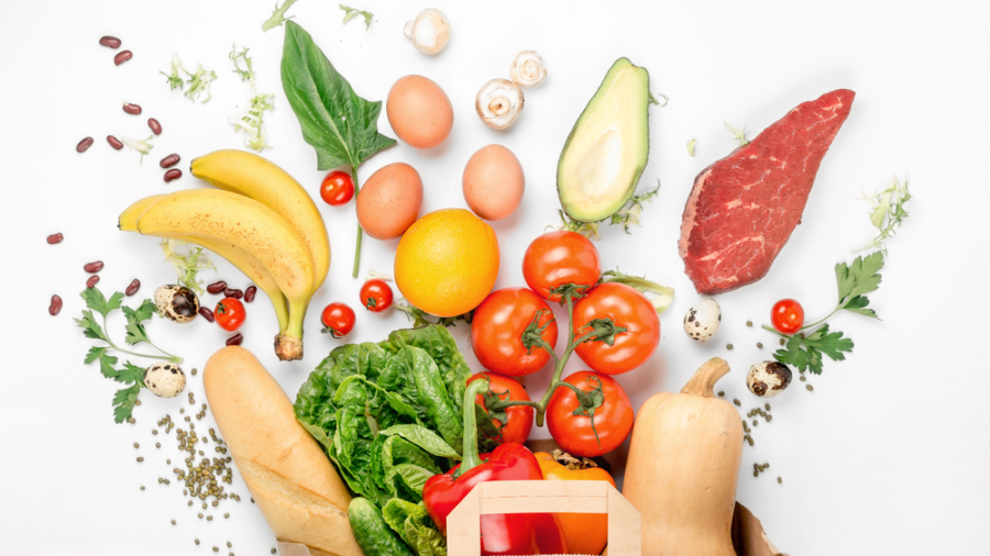 A paper bag filled with fresh vegetables, fruits, bread, and eggs is surrounded by assorted foods—each showcasing supply chain traceability—including bananas, tomatoes, spinach, steak, avocado, oranges, mushrooms, and quail eggs on a white background.