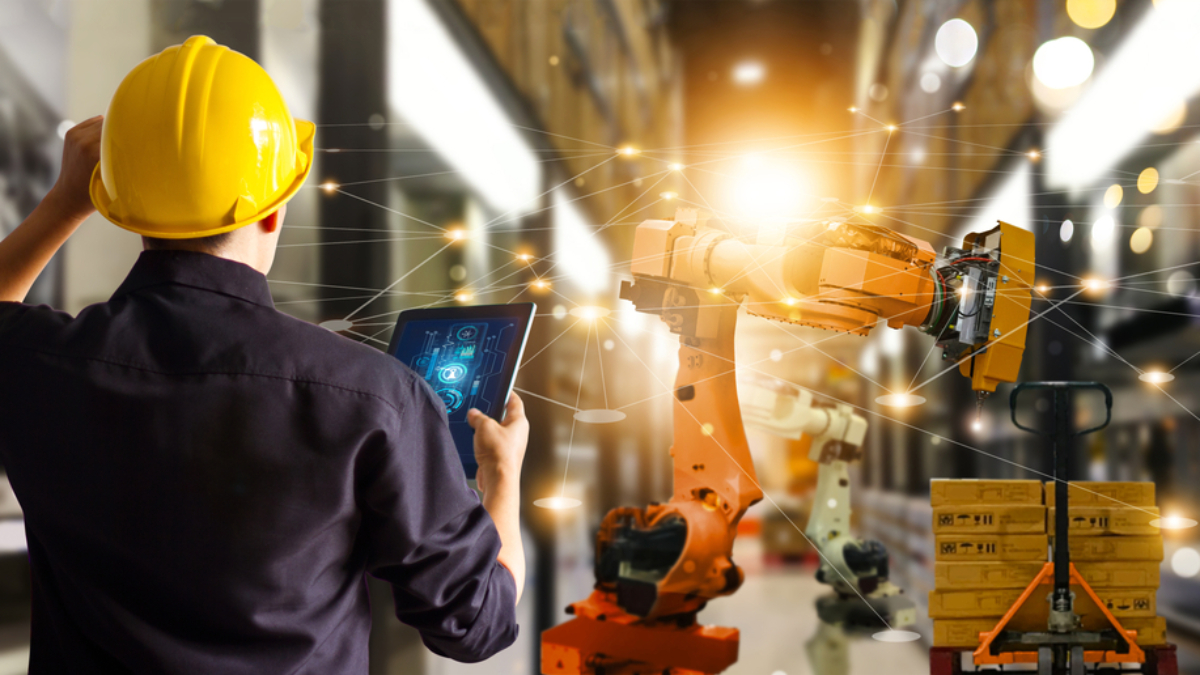 A person in a yellow hard hat uses a tablet to control an industrial robotics arm in a modern warehouse, with digital network lines connecting technology and equipment in the background.