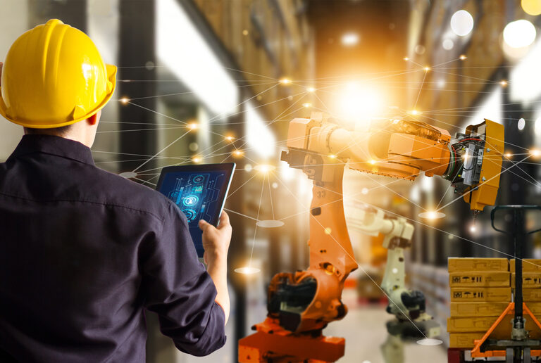 A person in a yellow hard hat uses a tablet to control an industrial robotics arm in a modern warehouse, with digital network lines connecting technology and equipment in the background.