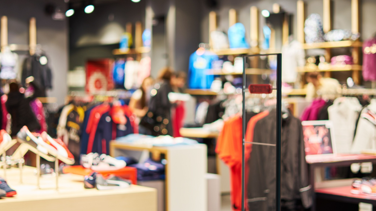 A brightly lit clothing and shoe store with various racks of colorful apparel and sneakers on display, shelves in the background, and a few customers browsing the merchandise.