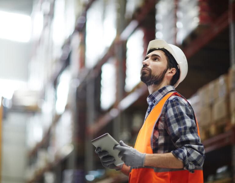 A warehouse worker wearing a hard hat, orange safety vest, gloves, and a plaid shirt uses a tablet for project management as he inspects shelves. The background shows stacked boxes on high racks.