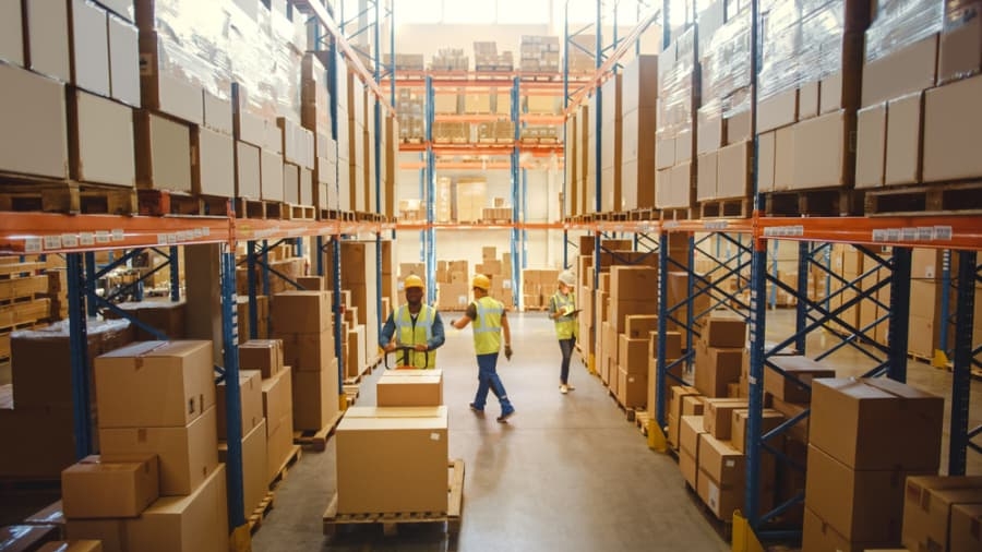 Warehouse workers in safety vests walk and talk between tall shelves filled with stacked boxes and packages during a Distribution Center Operational Assessment in a brightly lit warehouse.