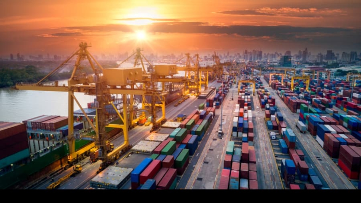 A busy shipping port at sunset with large cranes loading and unloading colorful shipping containers; city skyline is visible in the background.