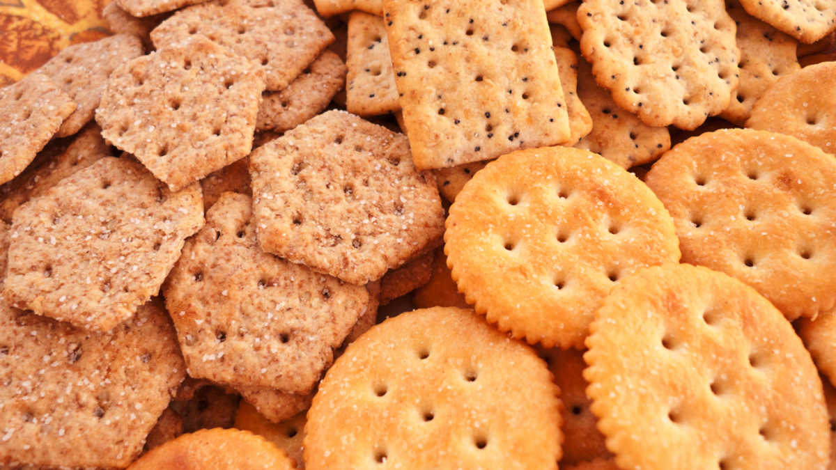 An assortment of different crackers, including hexagonal wheat crackers, square seeded crackers, and round buttery crackers, arranged close together on a patterned surface.