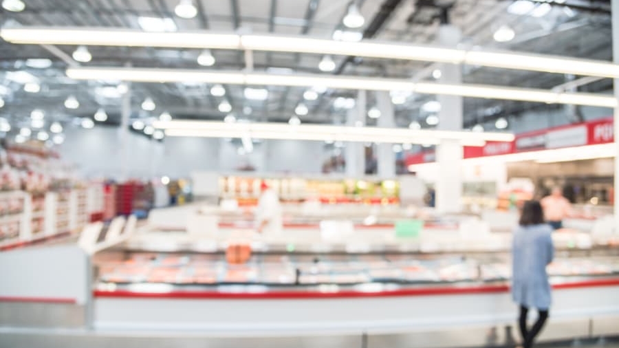 A blurred image of a supermarket interior with bright overhead lights, aisles, and display cases. A person stands near the counter in the foreground, but all details are out of focus.