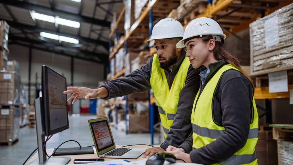 Two warehouse workers in safety vests and hard hats review order accuracy on a computer monitor in a storage facility, surrounded by shelves with stacked boxes and paperwork on a desk.