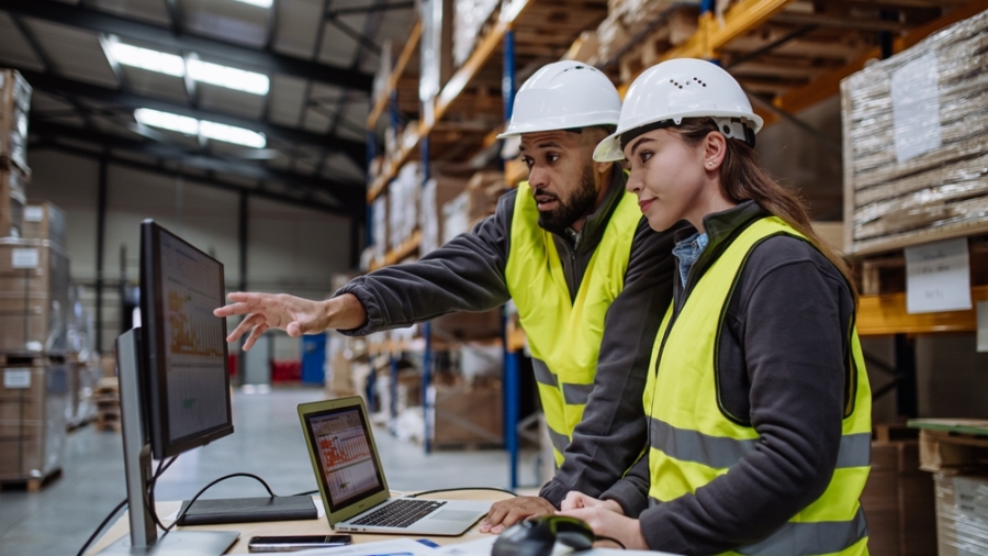 Two warehouse workers in safety vests and hard hats review order accuracy on a computer monitor in a storage facility, surrounded by shelves with stacked boxes and paperwork on a desk.