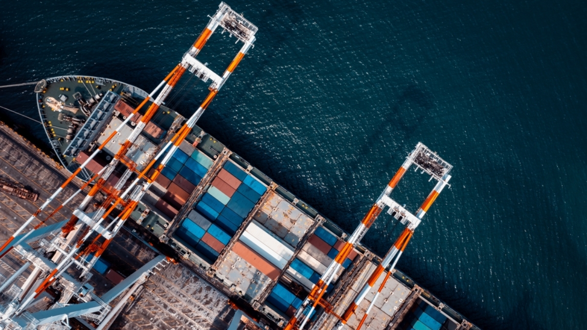 Aerial view of a cargo ship docked at a port, with colorful shipping containers and large orange-and-white cranes unloading goods—resembling the organized structure of Microsoft Fabric by the water.