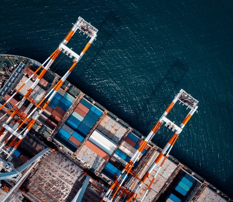 Aerial view of a cargo ship docked at a port, with colorful shipping containers and large orange-and-white cranes unloading goods—resembling the organized structure of Microsoft Fabric by the water.