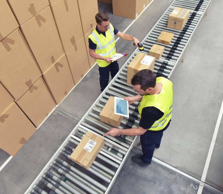 Two workforce members in yellow vests scan and check boxes on a conveyor belt in a warehouse. Stacks of cardboard boxes are visible nearby, and both workers are handling packages.