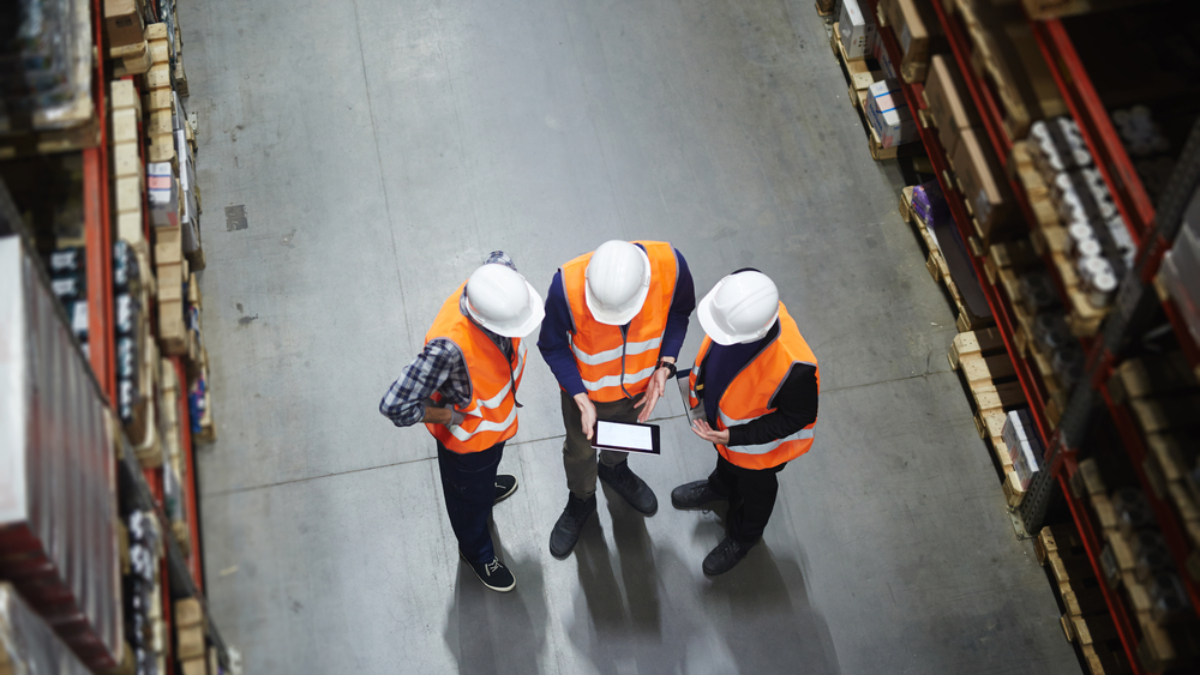 Three people in white hard hats and orange safety vests stand in a warehouse aisle, discussing what is supply chain strategy while looking at a tablet. Shelves filled with boxes line both sides of the concrete floor. View is from above.