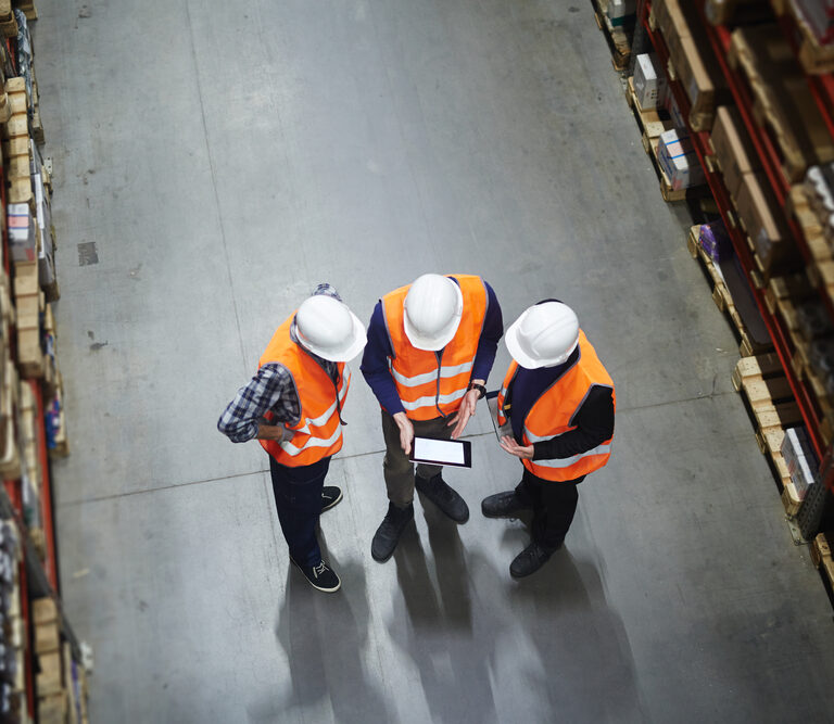 Three people in white hard hats and orange safety vests stand in a warehouse aisle, discussing what is supply chain strategy while looking at a tablet. Shelves filled with boxes line both sides of the concrete floor. View is from above.