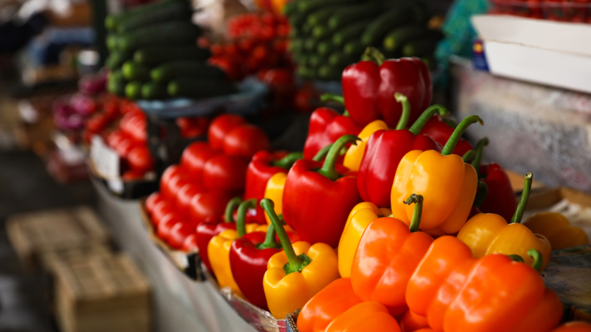 Colorful bell peppers—orange, yellow, and red—are displayed in rows at a market stall, with cucumbers and cherry tomatoes visible in the background, offering a vibrant scene reminiscent of cloud computing’s seamless organization.