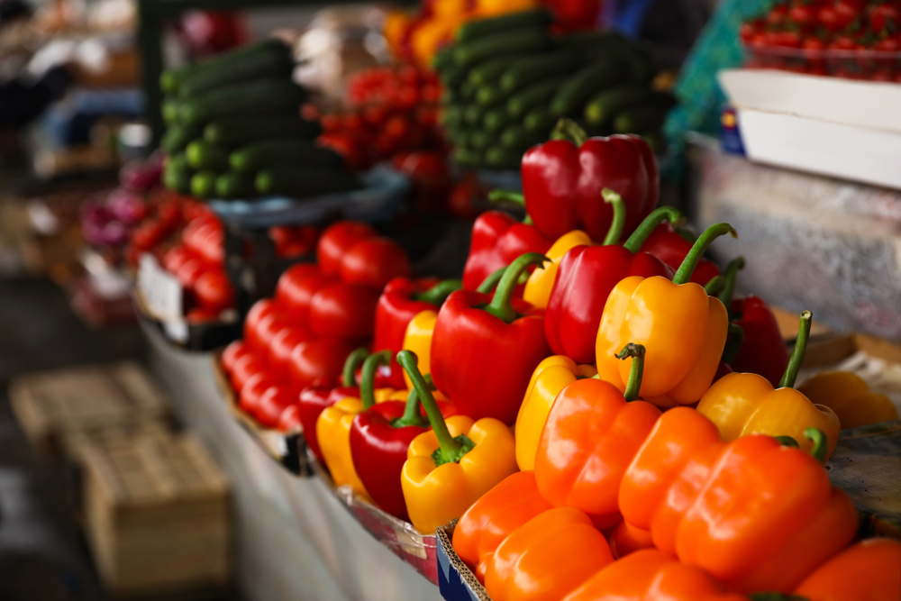 Colorful bell peppers—orange, yellow, and red—are displayed in rows at a market stall, with cucumbers and cherry tomatoes visible in the background, offering a vibrant scene reminiscent of cloud computing’s seamless organization.