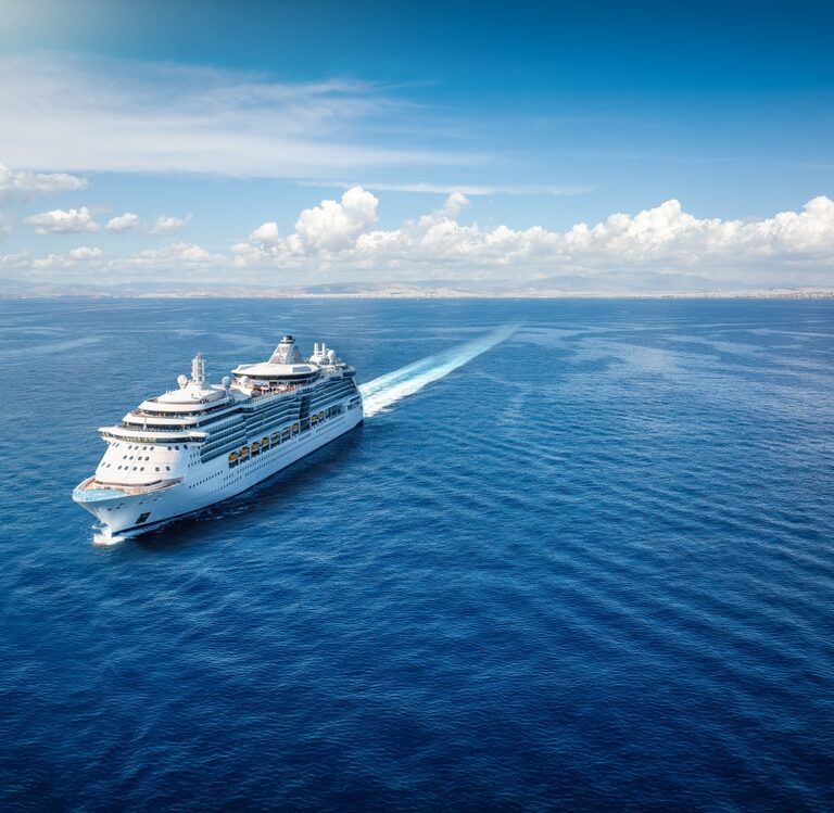 A large white cruise ship sails across a calm, deep blue sea under a bright sky with scattered clouds, leaving a wake behind it. Land is visible in the distance on the horizon.