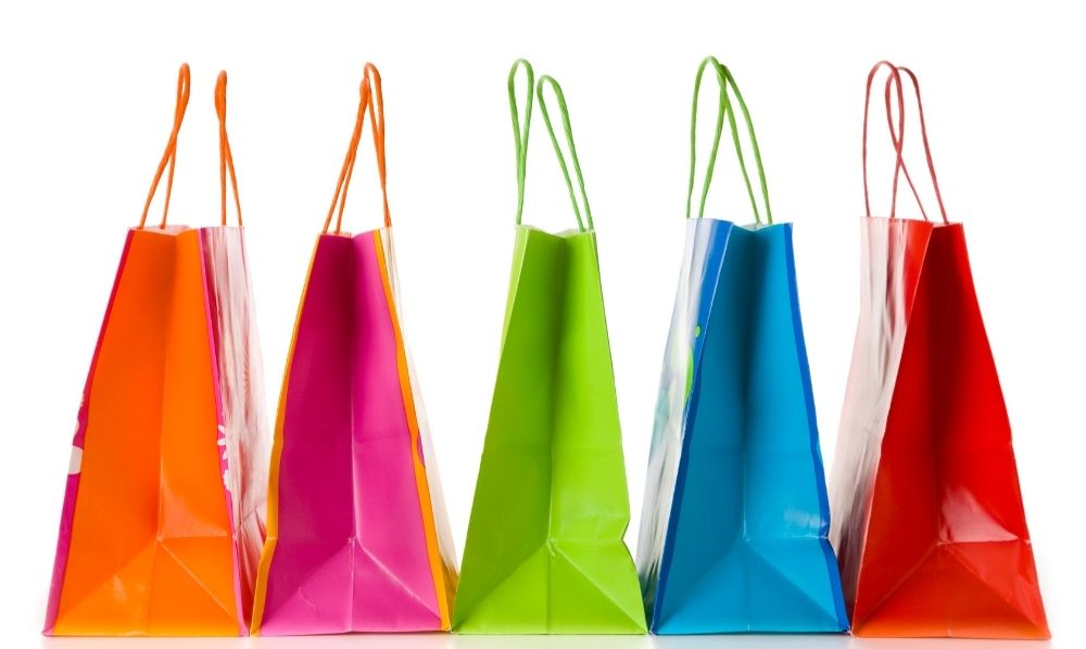 Five colorful shopping bags—orange, pink, green, blue, and red—are standing upright in a row against a white background, showcasing vibrant retail solutions with their handles clearly visible.
