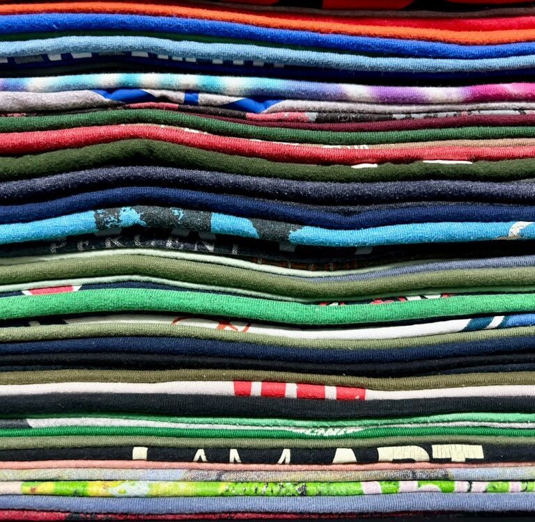 A close-up view of a neatly stacked pile of folded t-shirts in various colors and patterns, creating a layered, striped appearance.