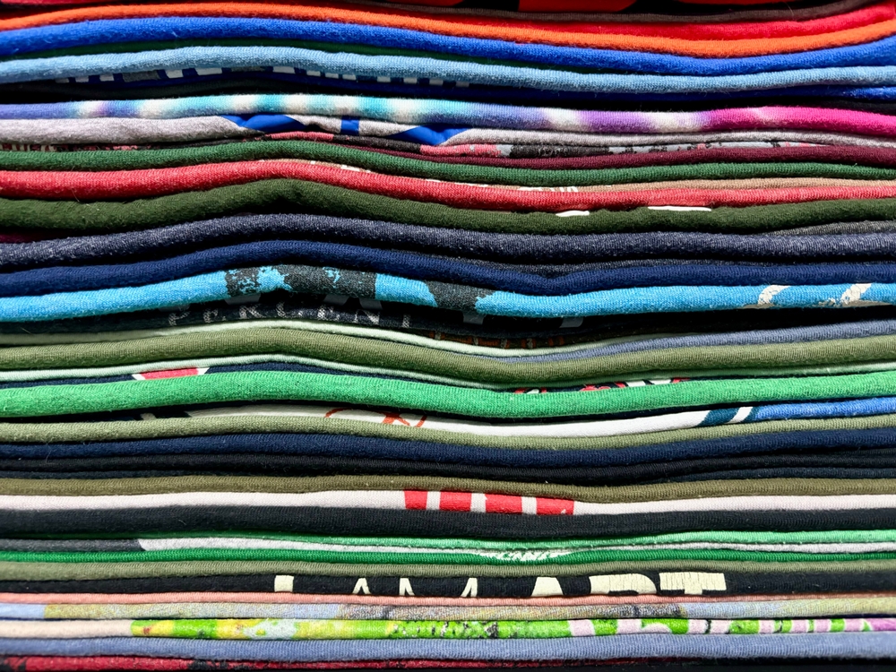 A close-up view of a neatly stacked pile of folded t-shirts in various colors and patterns, creating a layered, striped appearance.