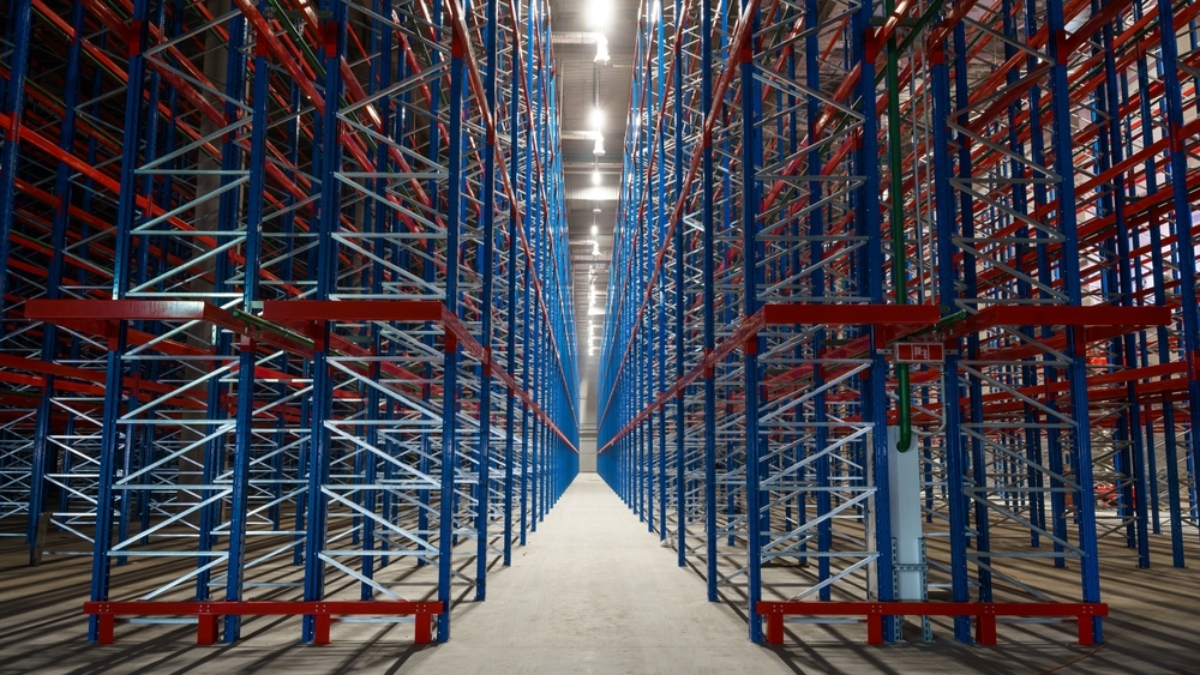 Empty metal storage racks extend in symmetrical rows inside a large warehouse, creating a sense of depth and perspective—an ideal setting for ai-powered inventory systems. Bright lights illuminate the clean concrete floor and steel framework.