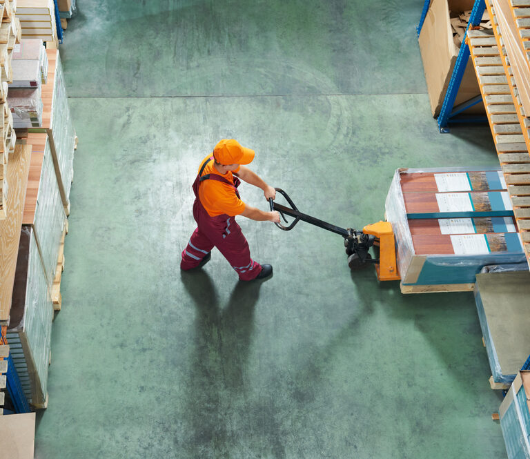A workforce member in an orange shirt and cap uses a pallet jack to move boxed goods between shelves on a green concrete floor, viewed from above.