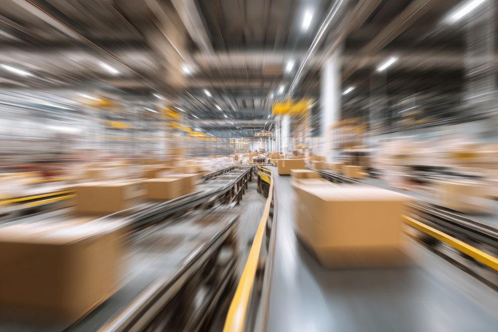 Blurred image of a busy warehouse or distribution center with many cardboard boxes moving quickly on conveyor belts, highlighting fast-paced logistics, shipping activity, and the importance of an efficient parcel audit service.