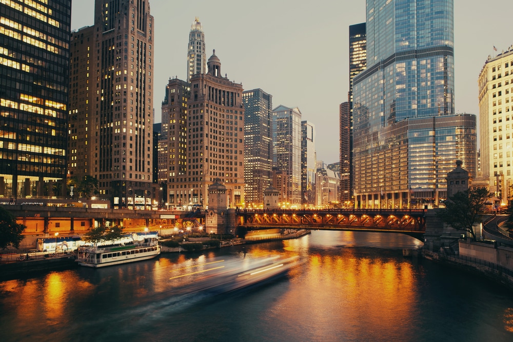 A cityscape at dusk showcases tall buildings and skyscrapers along the Chicago River, with a lit bridge and blurred boats moving on the water—reflecting city lights and enVista Careers' vibrant energy.
