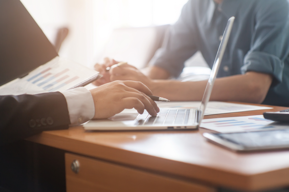 Two people sit at a desk with a laptop, papers, and charts, discussing documents and data—possibly exploring enVista Careers or strategies for their parcel audit service—in a brightly lit office.