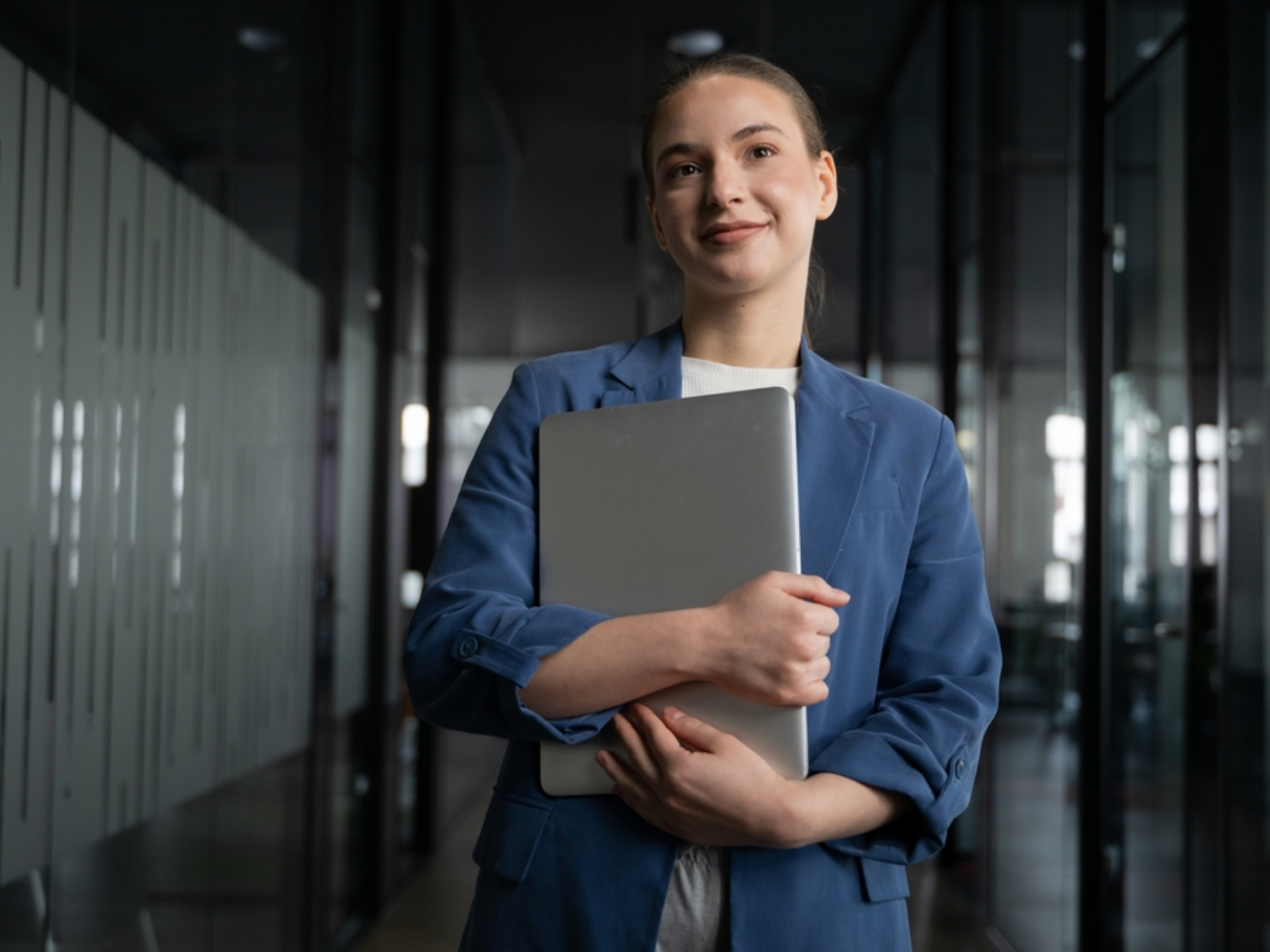 A woman wearing a blue blazer stands in a modern office hallway, smiling and holding a closed laptop to her chest—showcasing the professional environment of enVista Careers. Glass walls and a dark, sleek setting are visible in the background.