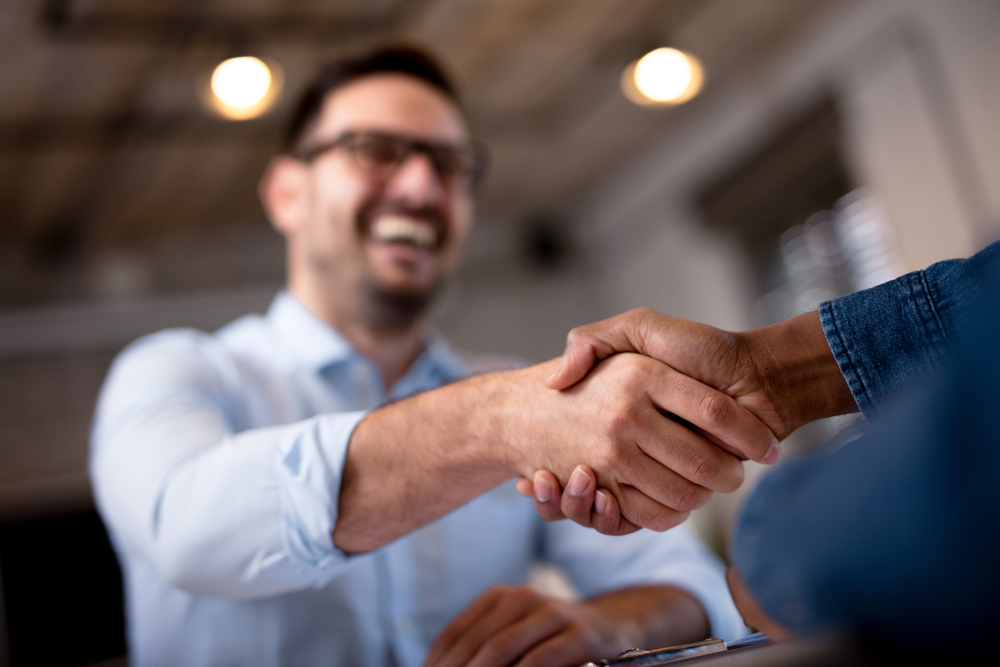 Two people shaking hands in a close-up shot, with one smiling man in the background wearing glasses and a light blue shirt, suggesting a successful enVista Careers meeting or friendly interaction about parcel audit service.