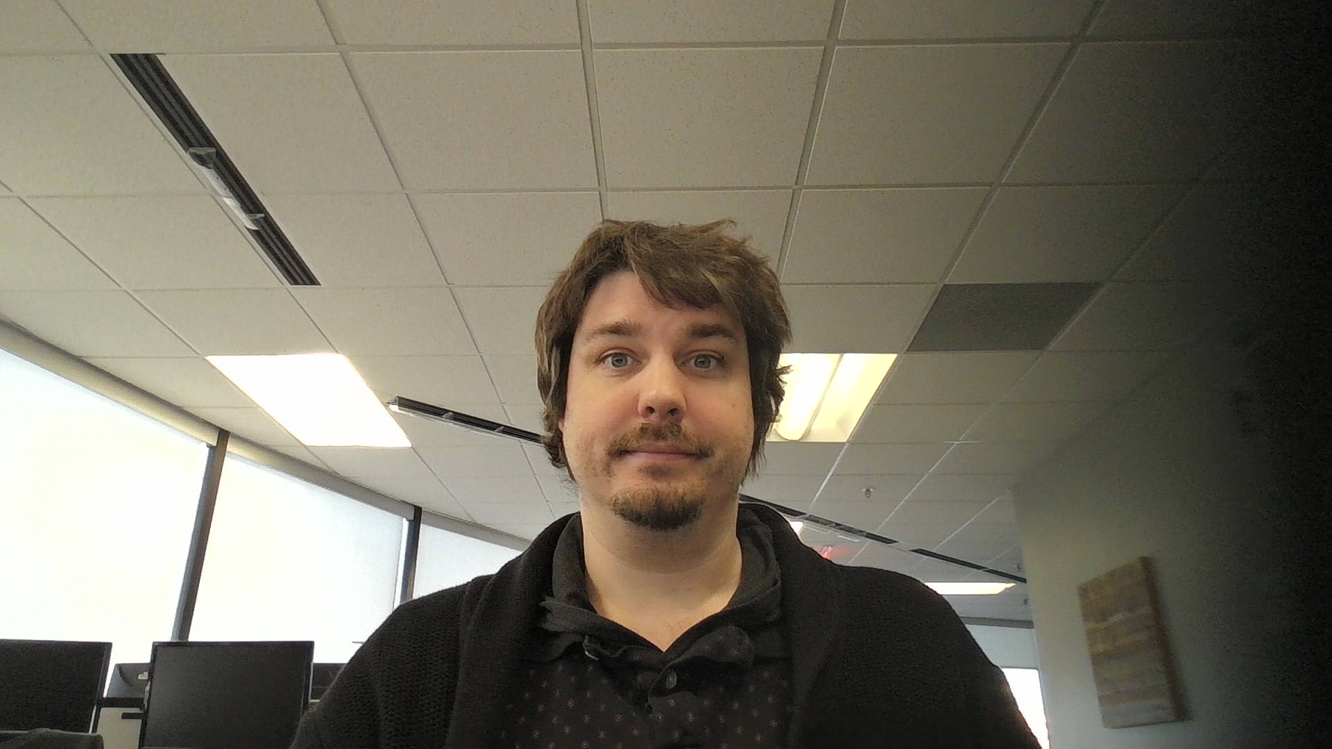 A man with light skin and brown hair is sitting indoors in an enVista Careers office with ceiling lights and large windows. He is looking at the camera with a neutral expression, surrounded by desks and computer monitors, possibly discussing parcel audit service.