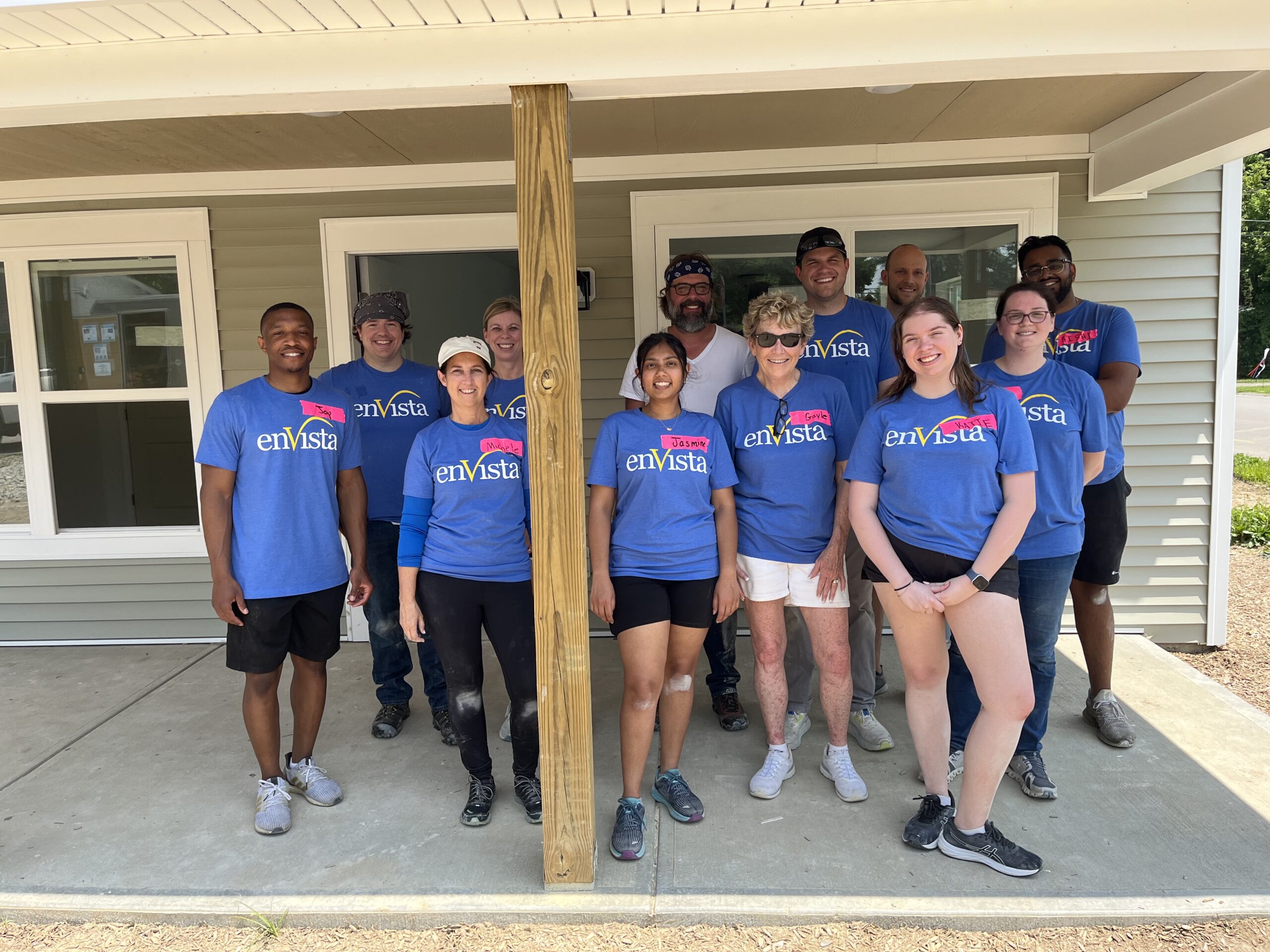A group of twelve people wearing matching blue enVista shirts, specialists in inventory optimization consulting, stand and smile together outside a light green house with white trim, posing for a group photo on a sunny day.