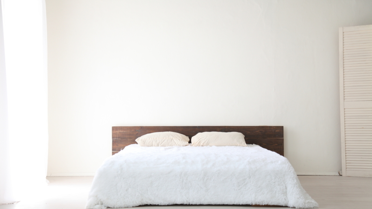 A minimalist bedroom with a low wooden headboard, two beige pillows, and a white fluffy comforter on the bed. White walls and a light carpet reflect pick-to-light levels of natural brightness throughout the space.