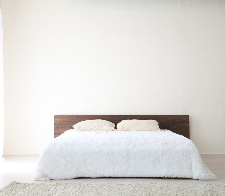 A minimalist bedroom with a low wooden headboard, two beige pillows, and a white fluffy comforter on the bed. White walls and a light carpet reflect pick-to-light levels of natural brightness throughout the space.
