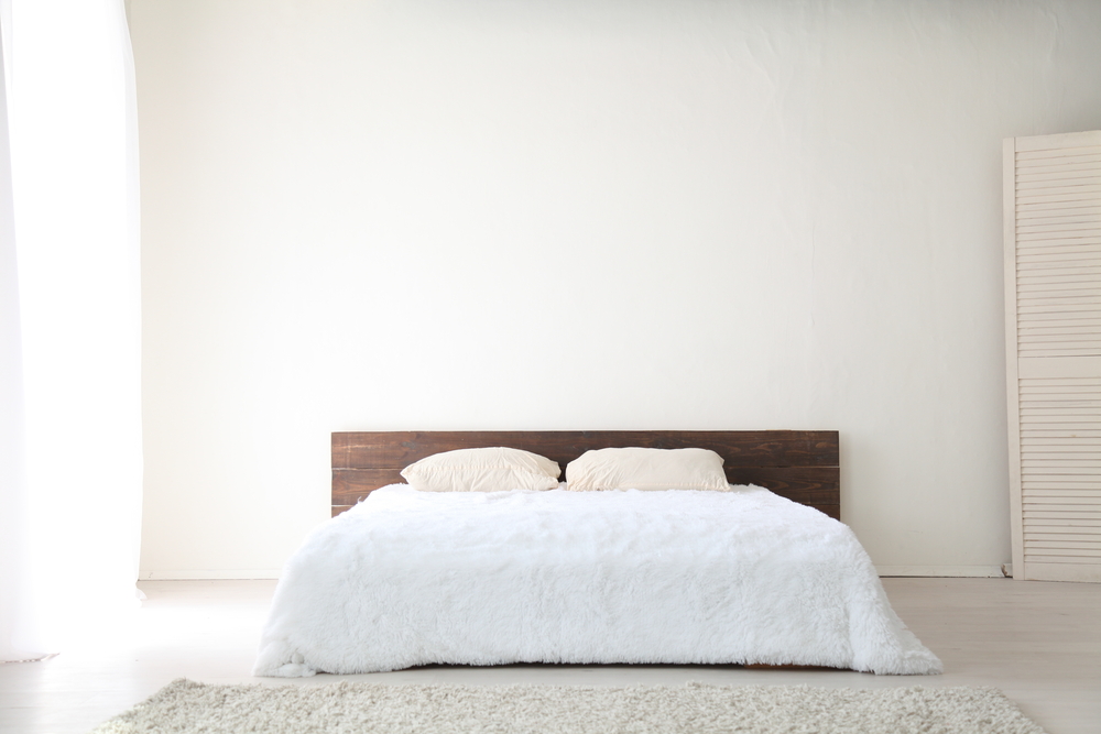 A minimalist bedroom with a low wooden headboard, two beige pillows, and a white fluffy comforter on the bed. White walls and a light carpet reflect pick-to-light levels of natural brightness throughout the space.