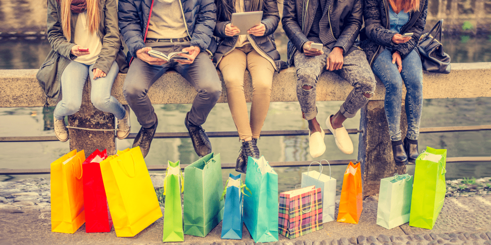 Five people sit on a stone bench outdoors, each with shopping bags of various colors and patterns placed in front of them. They are dressed in casual winter clothes and appear to be using their phones or reading.