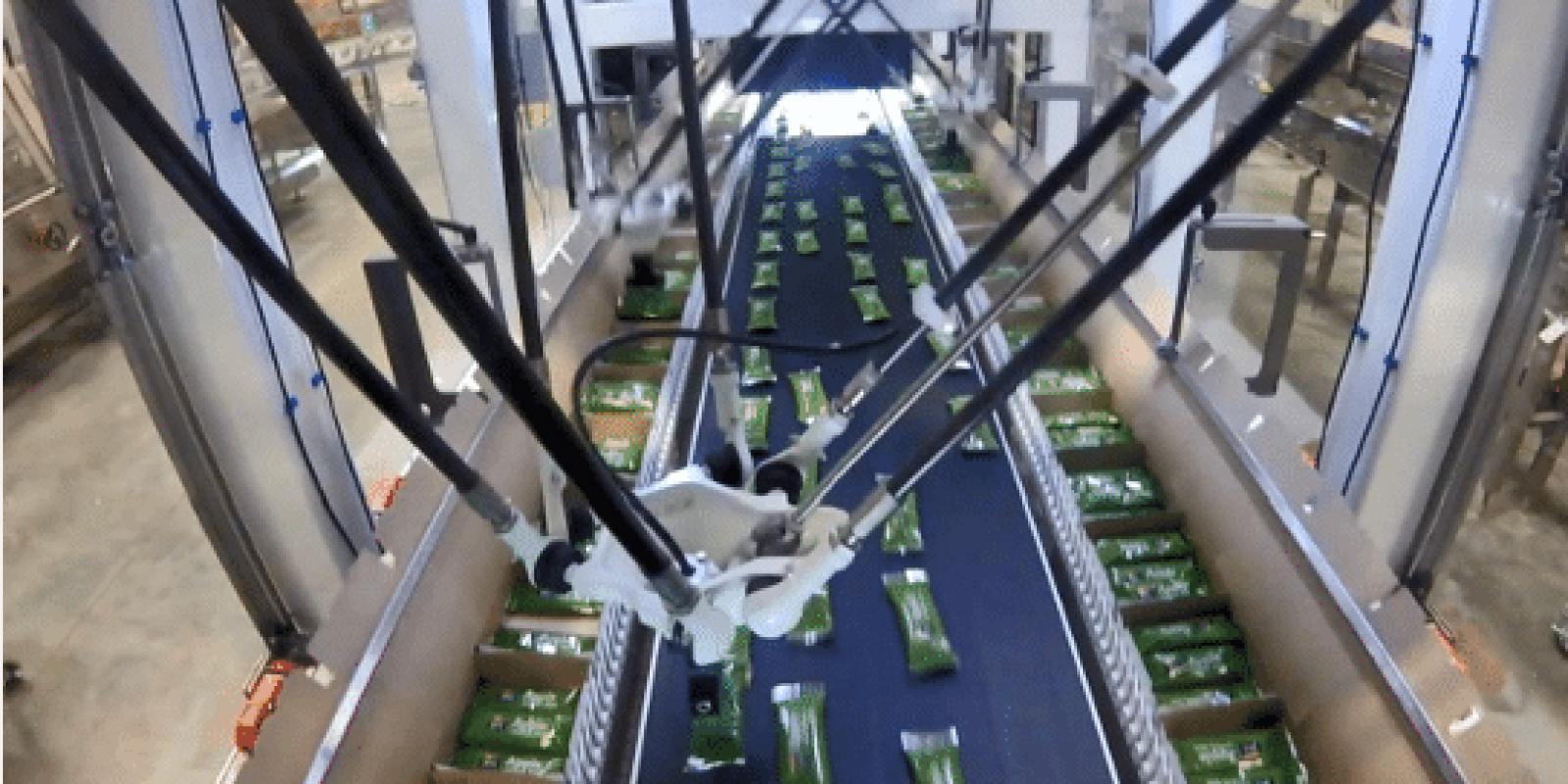 Overhead view of an automated robotic arm sorting green rectangular packages on a conveyor belt inside a large industrial facility.
