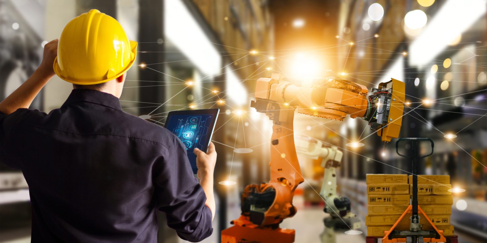 A person in a yellow hard hat uses a tablet to control an industrial robotics arm in a modern warehouse, with digital network lines connecting technology and equipment in the background.