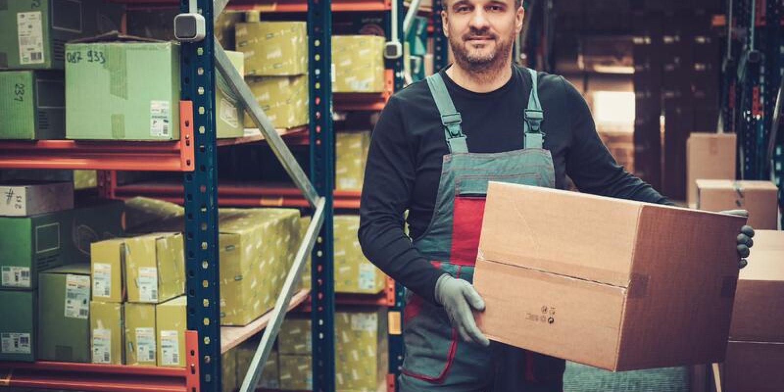 A warehouse worker in overalls and gloves holds a cardboard box, standing among shelves filled with packages and parcels. The storage area is organized and well-lit.