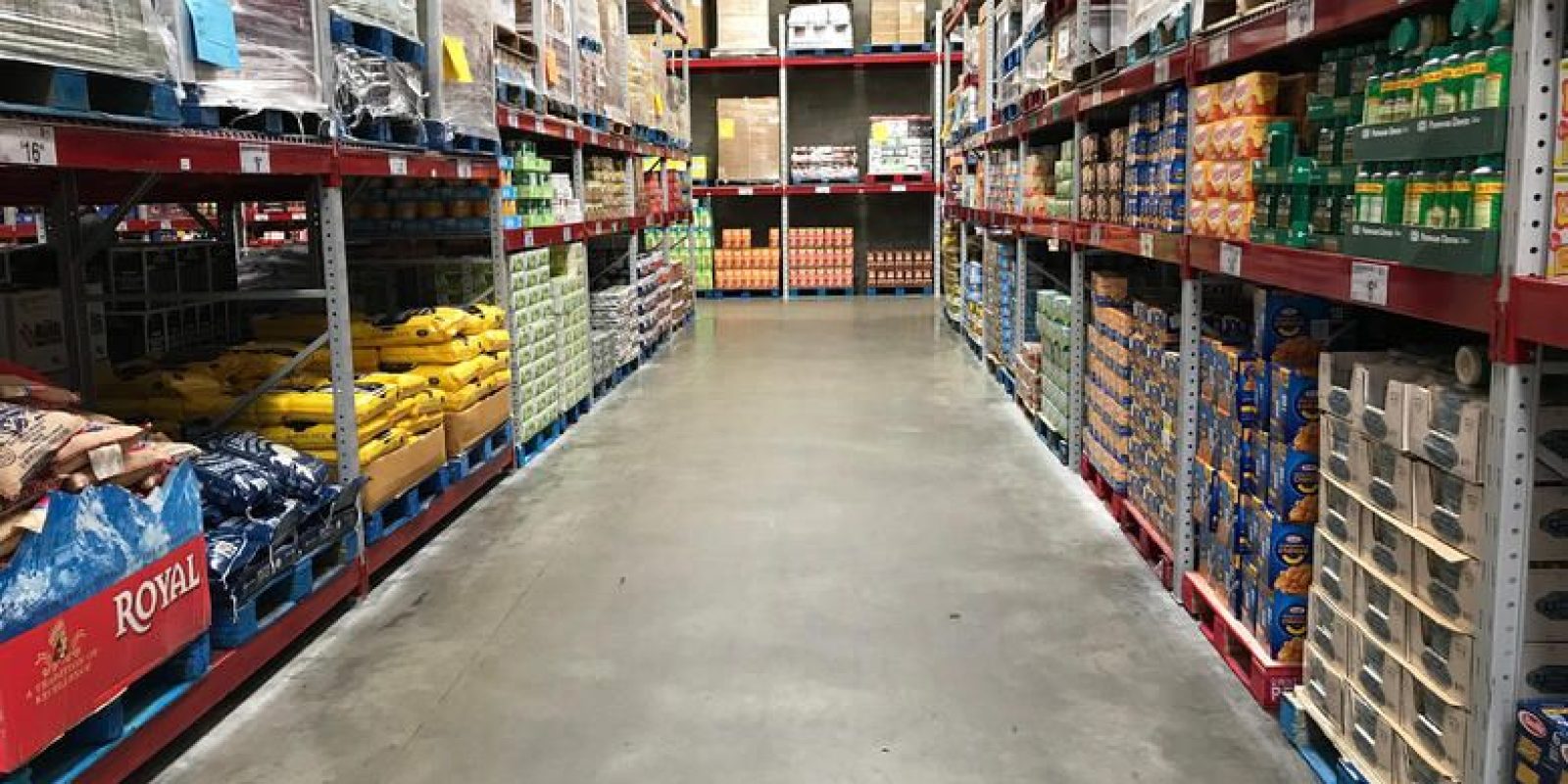 A wide aisle in a warehouse-style grocery store with shelves stacked high with bulk food items and large boxes on both sides. The concrete floor is clean and there are no shoppers visible.