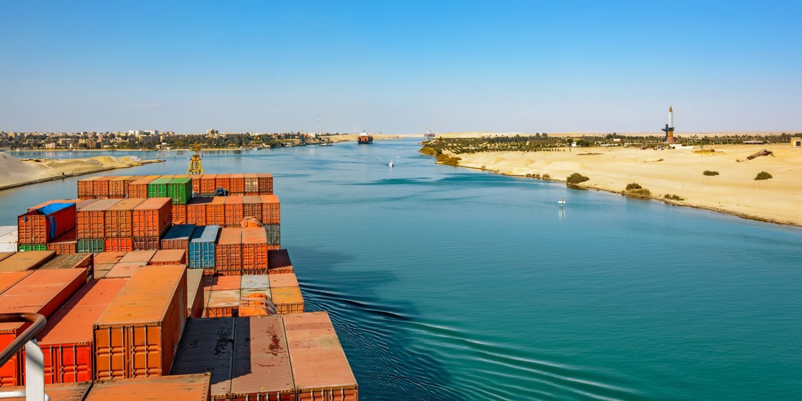 A cargo ship loaded with containers sails through the Suez Canal, bordered by sandy desert on one side and a distant cityscape on the other, under a clear blue sky.