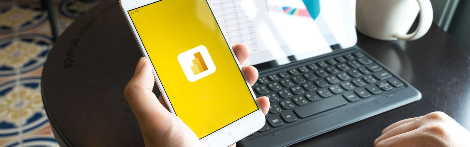 A person holds a smartphone displaying the Microsoft Power BI logo, while sitting at a round table with a tablet showing a colorful pie chart and a white coffee mug.