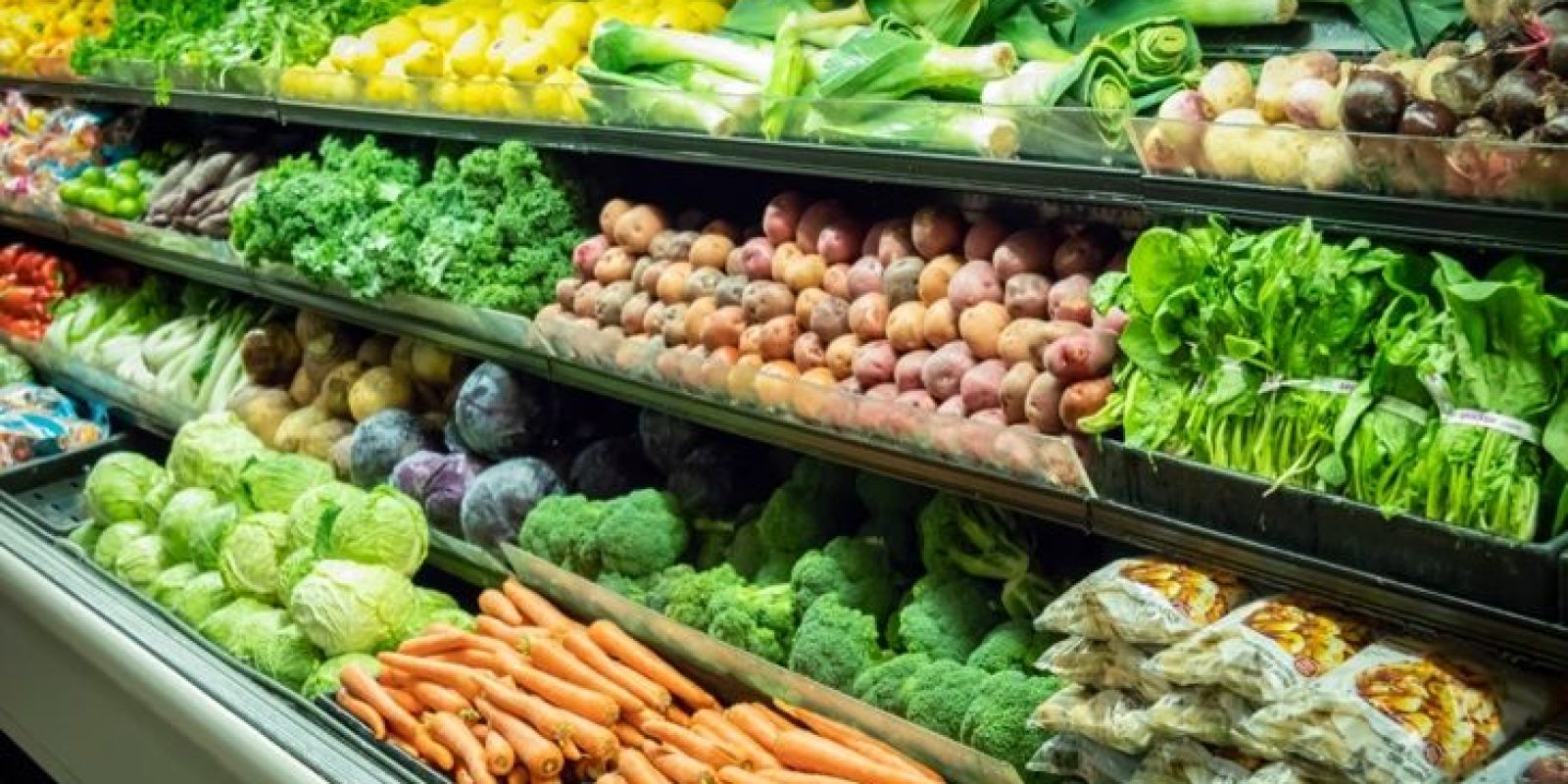 A grocery store produce section displaying a variety of fresh vegetables, including carrots, lettuce, broccoli, potatoes, leeks, bell peppers, and leafy greens neatly arranged on shelves.