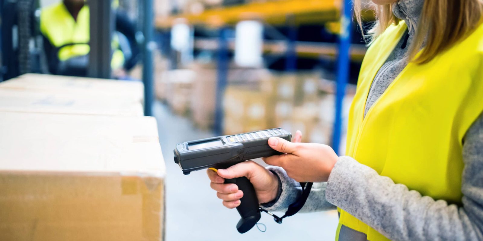 A worker in a yellow safety vest scans a cardboard box with a handheld barcode scanner in a warehouse. Shelves with more boxes and another worker in the background are visible.
