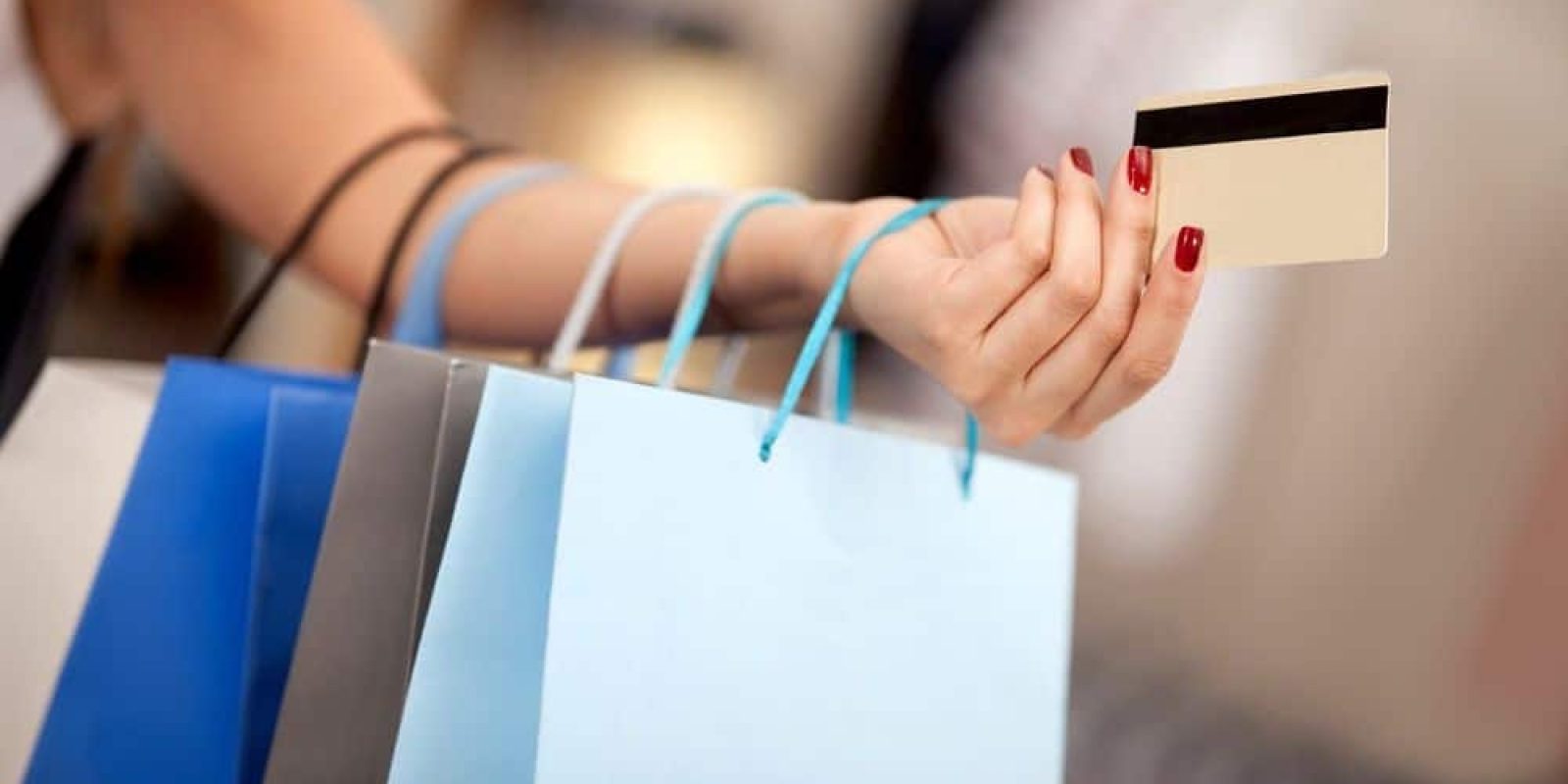 A person with red-painted nails holds several shopping bags and a credit card, suggesting a shopping activity. The background is blurred, keeping the focus on the hand, bags, and card.
